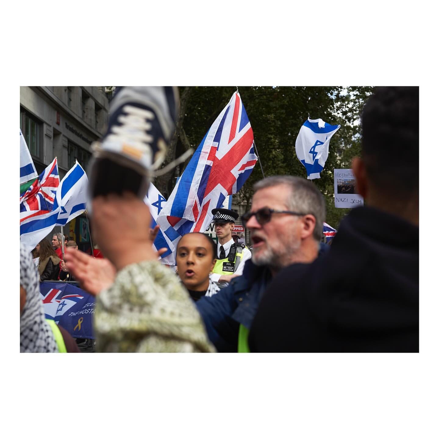 Crowd with UK and Israeli flags rallying during a protest in an urban environment with buildings, people, and a police officer.