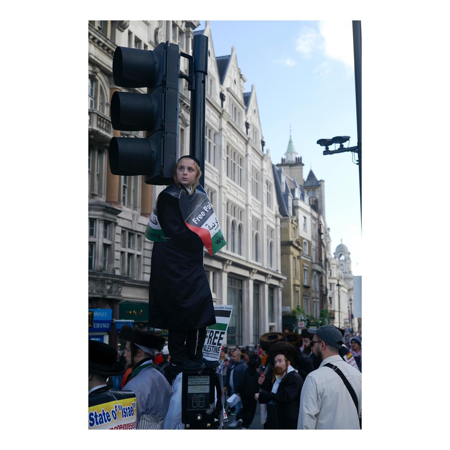 Woman stands on traffic light pole in urban setting, draped in flag with street crowd, buildings, and blue sky in background.