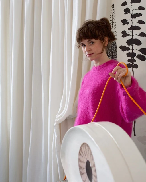 Woman in pink sweater poses indoors with white decorative item and orange cord against a backdrop of white curtains and tree art.