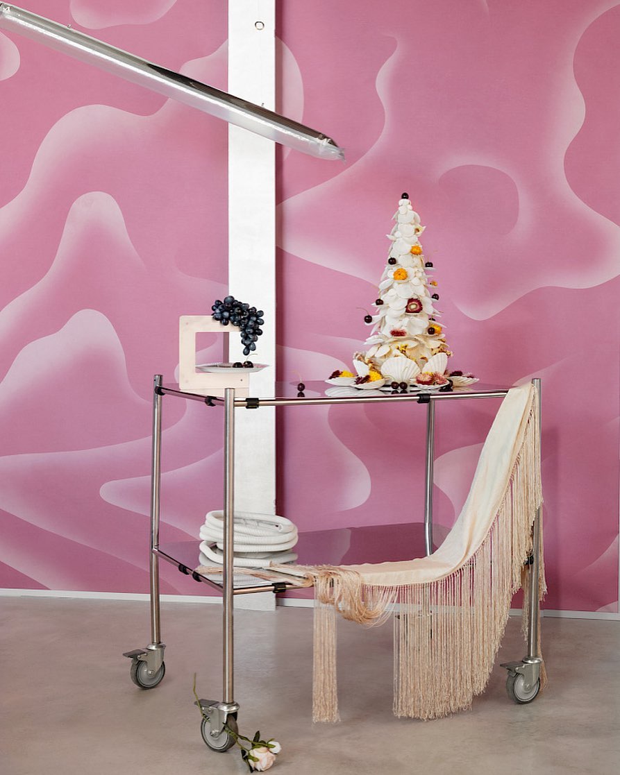 Towering dessert on a metal service cart against a pink patterned wall, featuring grapes, draped fabric, and a minimalist aesthetic.