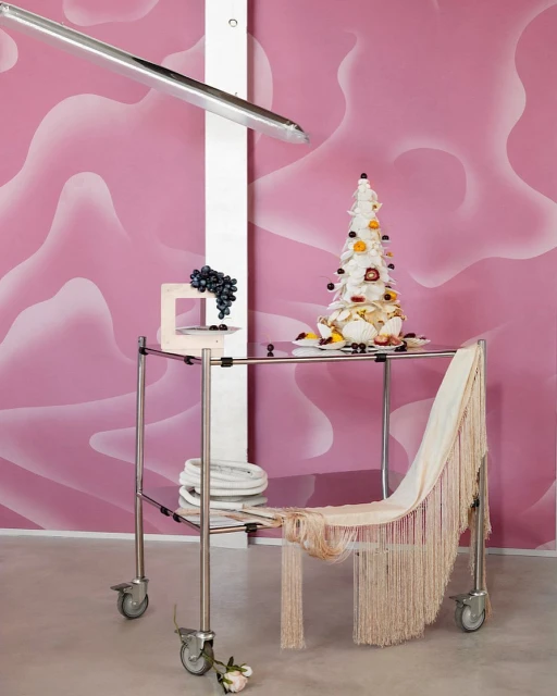 Towering dessert on a metal service cart against a pink patterned wall, featuring grapes, draped fabric, and a minimalist aesthetic.