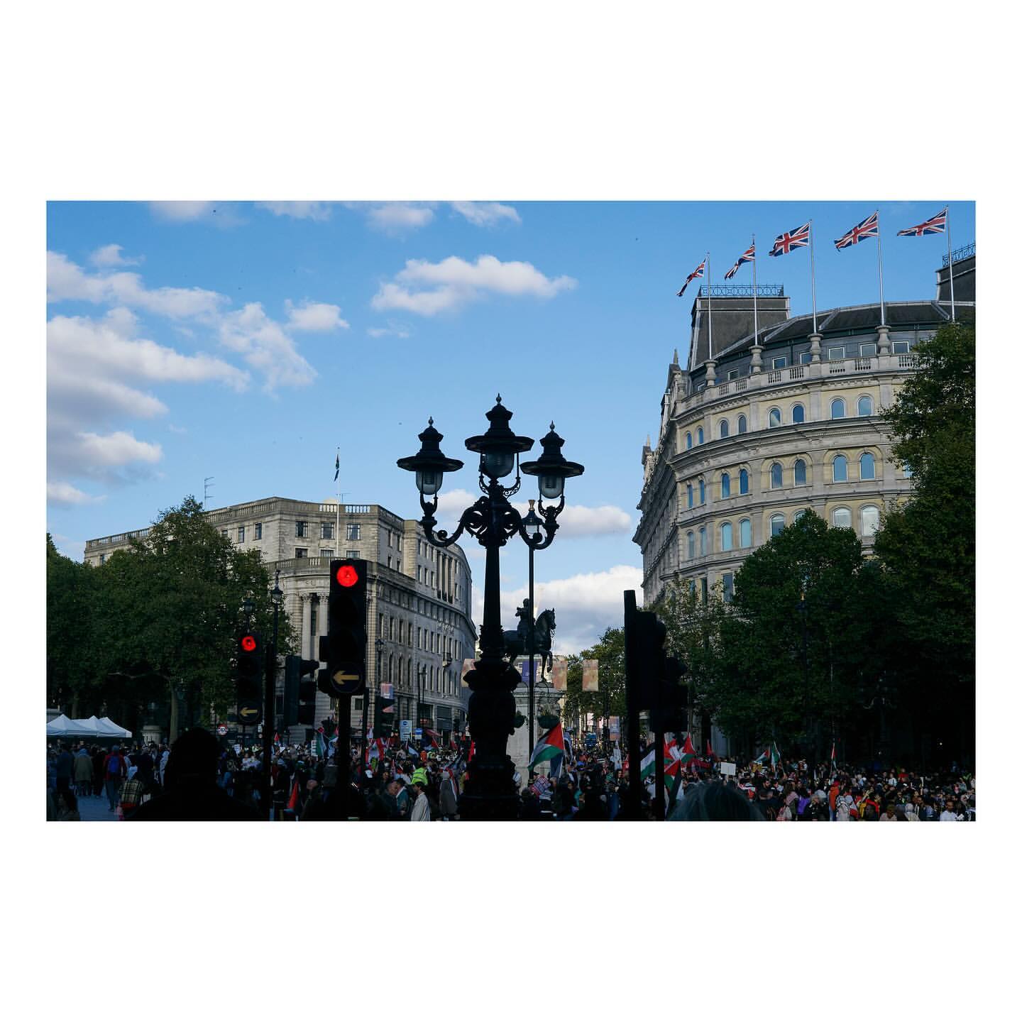 Crowd of people gather in a city square with flags and historic architecture under a partly cloudy sky; street lamp prominent.