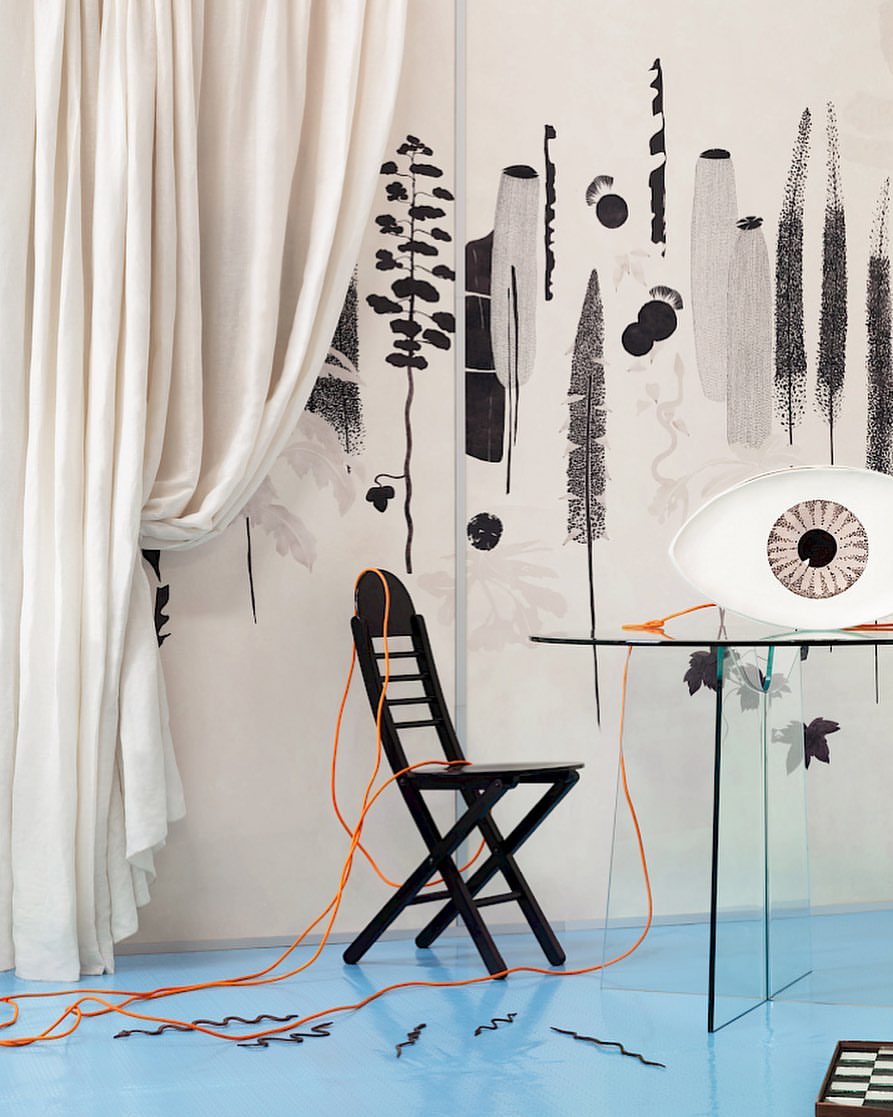 Interior shot with black folding chair, glass table, and 'eye' lamp against a backdrop of abstract nature-themed black-and-white art.