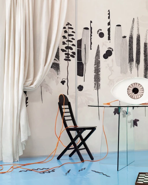 Interior shot with black folding chair, glass table, and 'eye' lamp against a backdrop of abstract nature-themed black-and-white art.