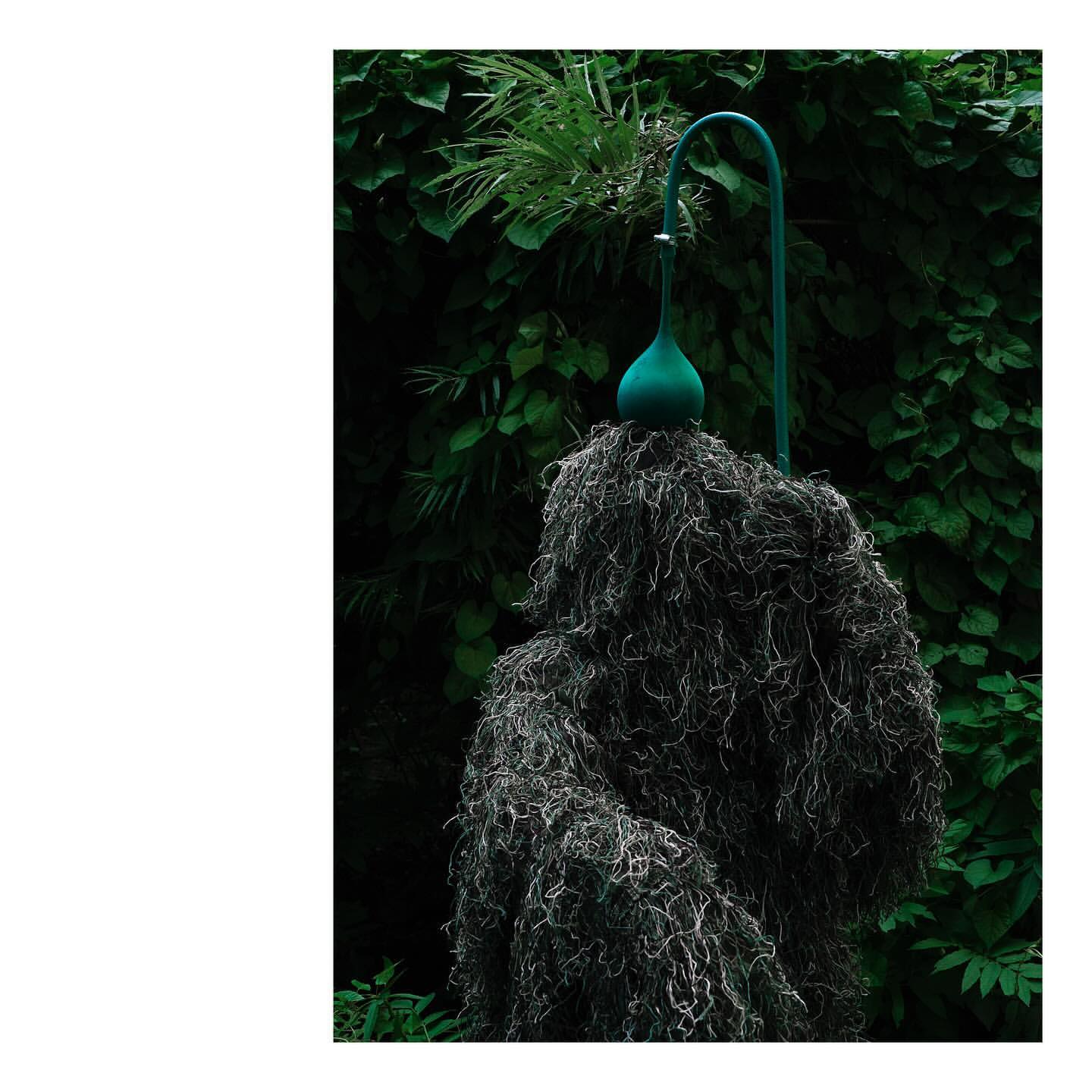 Moss-covered sculpture in garden with green gourd and metal support, set against a dark green leafy background, artistic presentation.