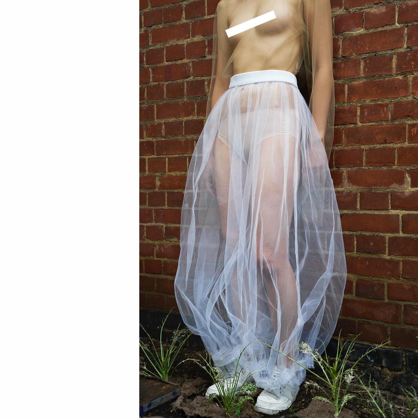 Adult female poses in a sheer skirt and top against a brick wall outdoors. She is wearing white sneakers and standing in front of plants.