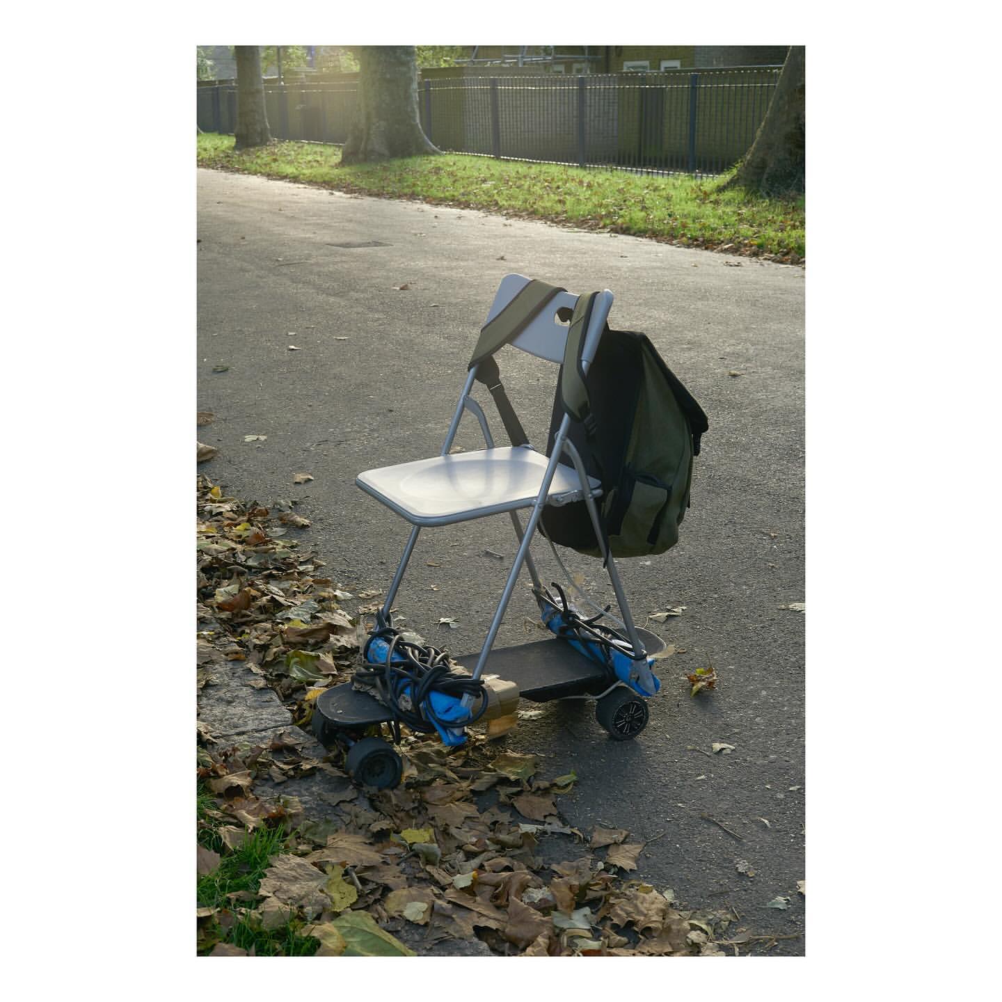 Chair mounted on a skateboard outside on a sidewalk with fallen leaves. A dark green backpack is hanging on the chair.