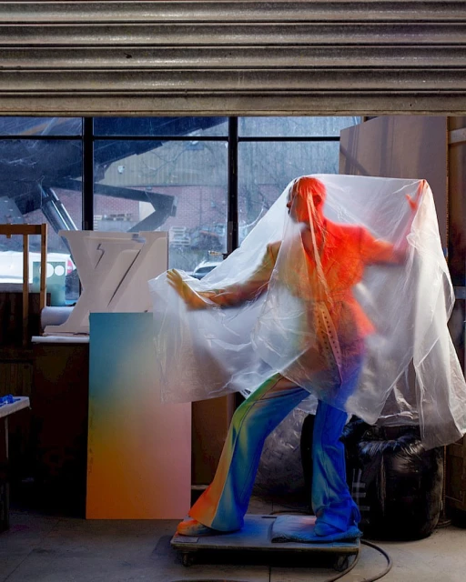 A person draped in translucent plastic stands in a studio, featuring colorful gradient artwork and a large window with a gray industrial door.