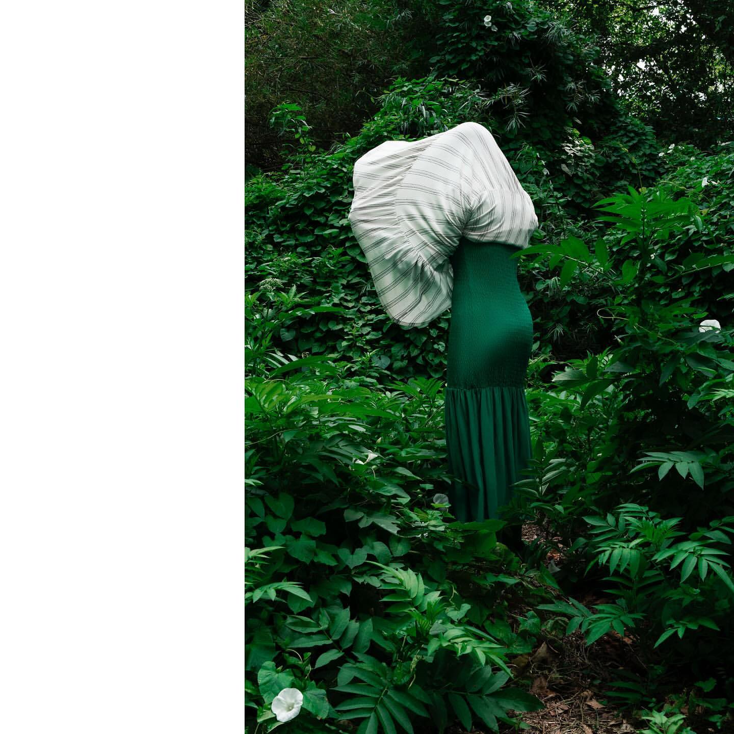 A woman in a green dress and striped top stands in a lush green forest, creating an abstract and nature-filled fashion image.