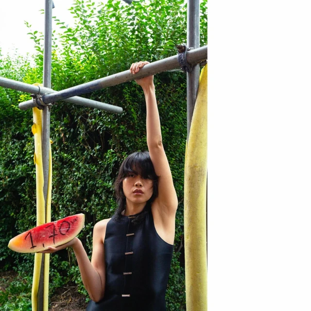 A young adult female poses holding a slice of watermelon while hanging from playground bars in an outdoor, natural setting.
