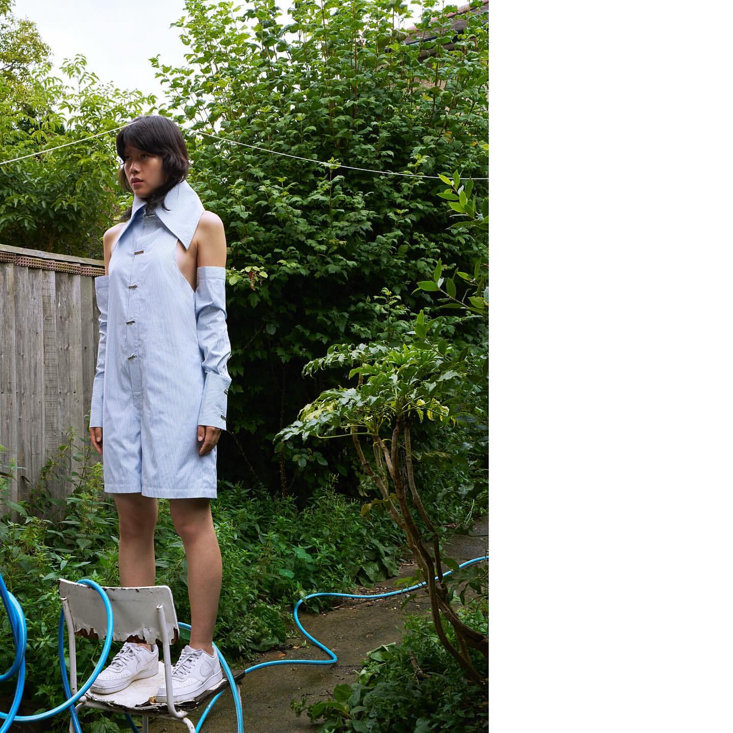 Woman in open-shoulder pinstripe romper stands on chair outside with green foliage, blue hose, and natural light.