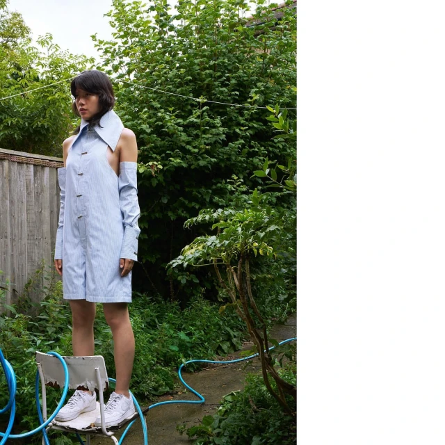 Woman in open-shoulder pinstripe romper stands on chair outside with green foliage, blue hose, and natural light.