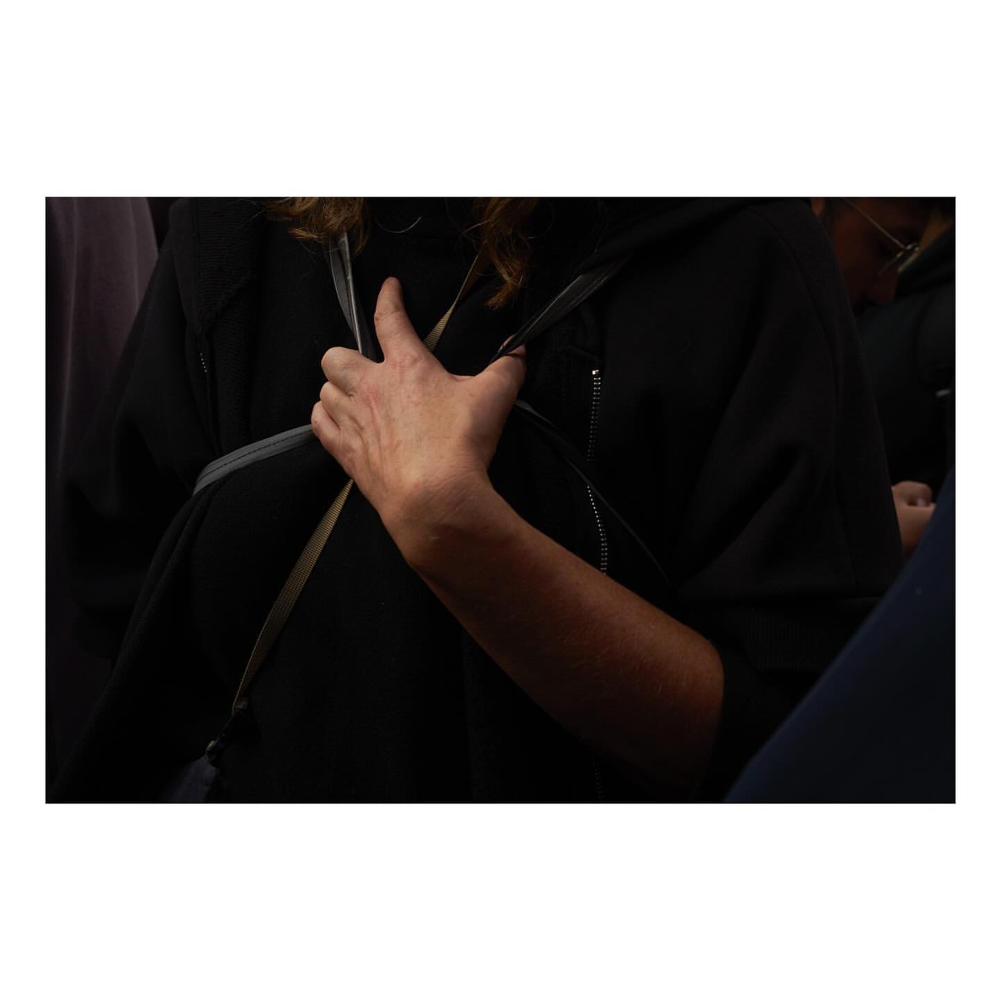 Close-up of a person wearing a black hooded jacket and backpack, holding the strap of the backpack with their hand. The shot is indoors.