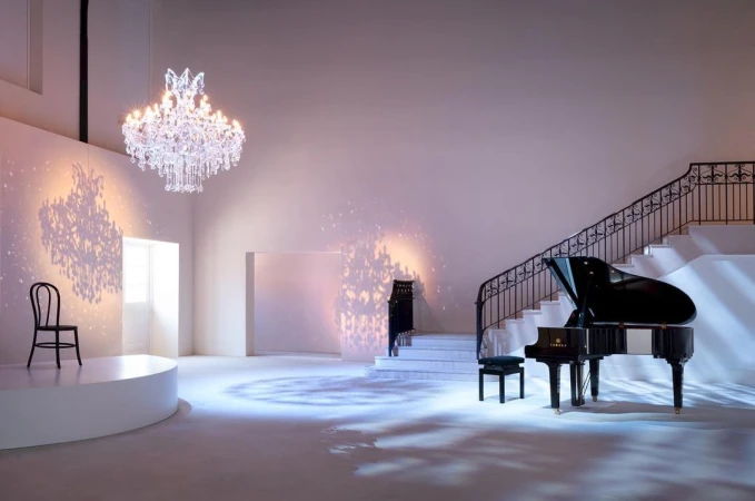 Interior of a spacious white room featuring a grand piano, elegant chandelier, chair on a platform, and a staircase with iron railings.