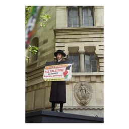 Man wearing a black coat and hat standing outside a stone building holding a political sign that reads Torah Demands All Palestine