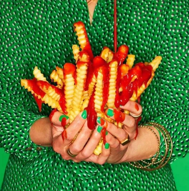 Close-up of hands holding french fries drizzled with ketchup against a vibrant green backdrop, showcasing a playful food composition.