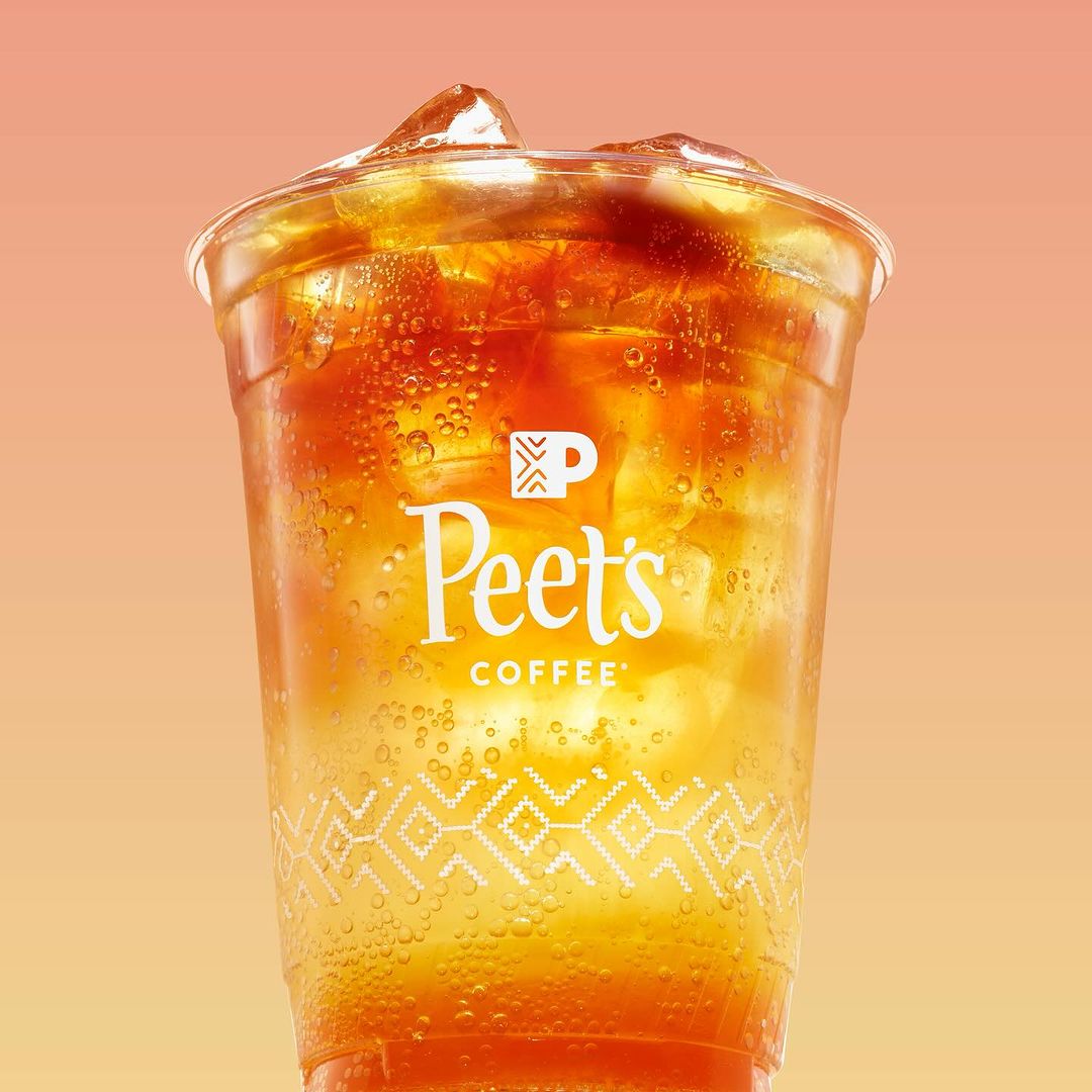 Iced coffee drink from Peet's Coffee in a clear plastic cup with logo and geometric design against an orange gradient background.