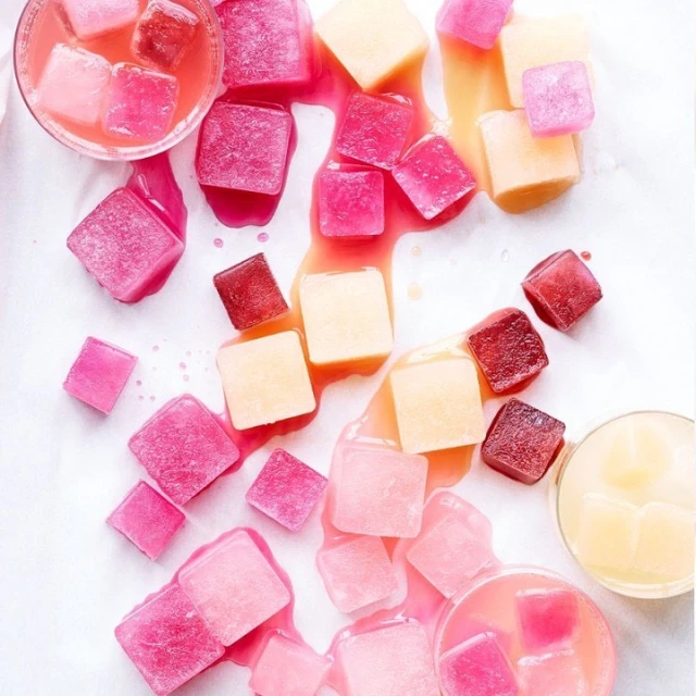 Overhead shot of colorful ice cubes of different hues on white background, scattered around glass cups filled with juice.