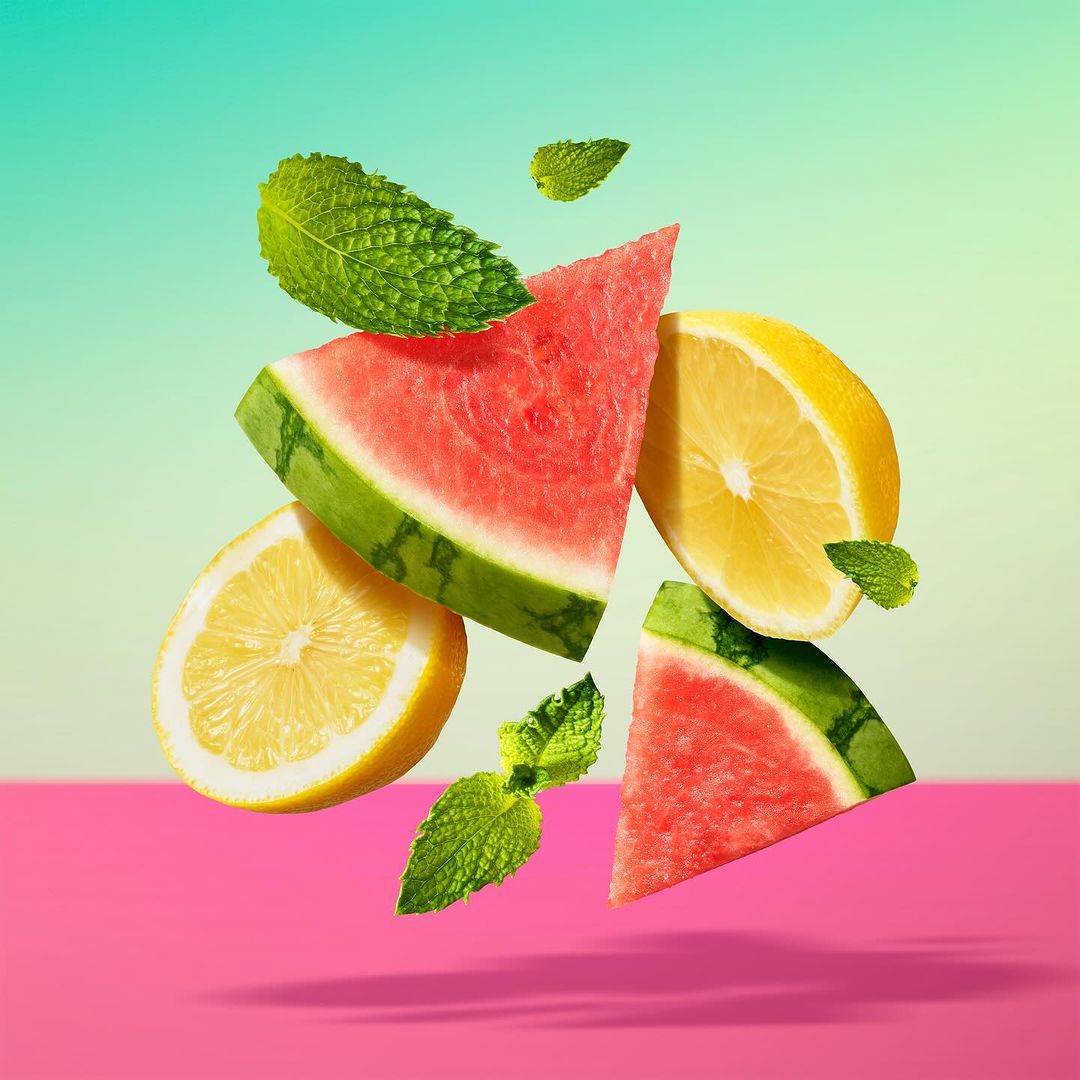 Slices of watermelon and lemons with mint leaves are floating in the air against a split pink and teal background.