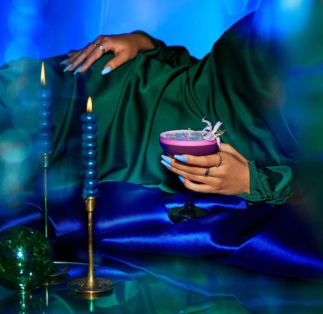 A hand holding a cocktail glass with a candle, adorned with a white flower, set against draped green and blue satin fabrics.