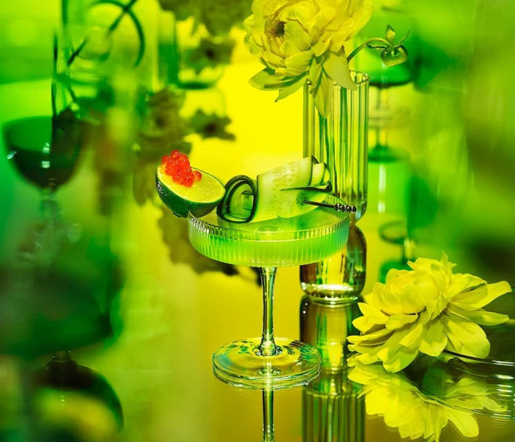 Stylized shot of a green cocktail in a stemmed glass with lime, cucumber, and red roe garnish, set against a green and yellow background.