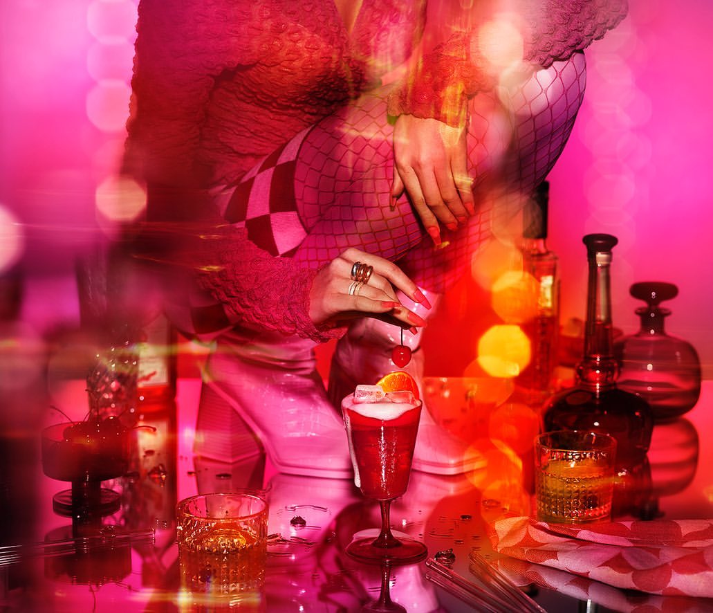 Woman in pink attire adds a cherry to a red cocktail. Bottles and glassware are on a mirrored surface, with a pink and red background.
