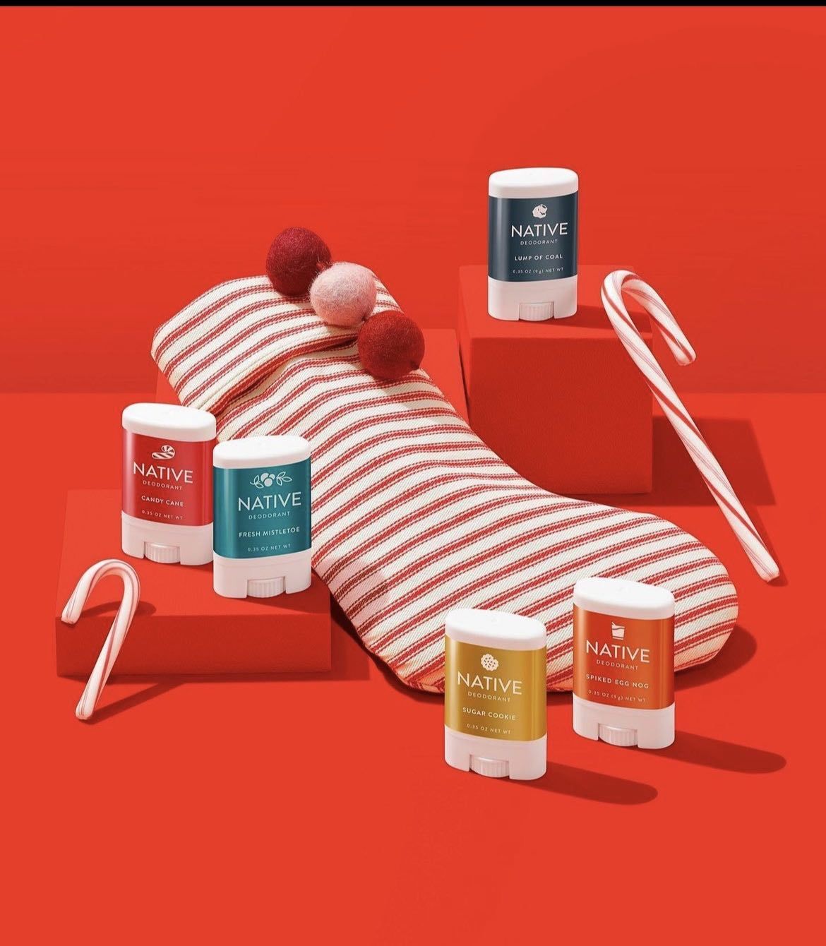 Festive holiday scene featuring an open Christmas stocking overflowing with assorted Native brand deodorants and two candy canes on a red backdrop.