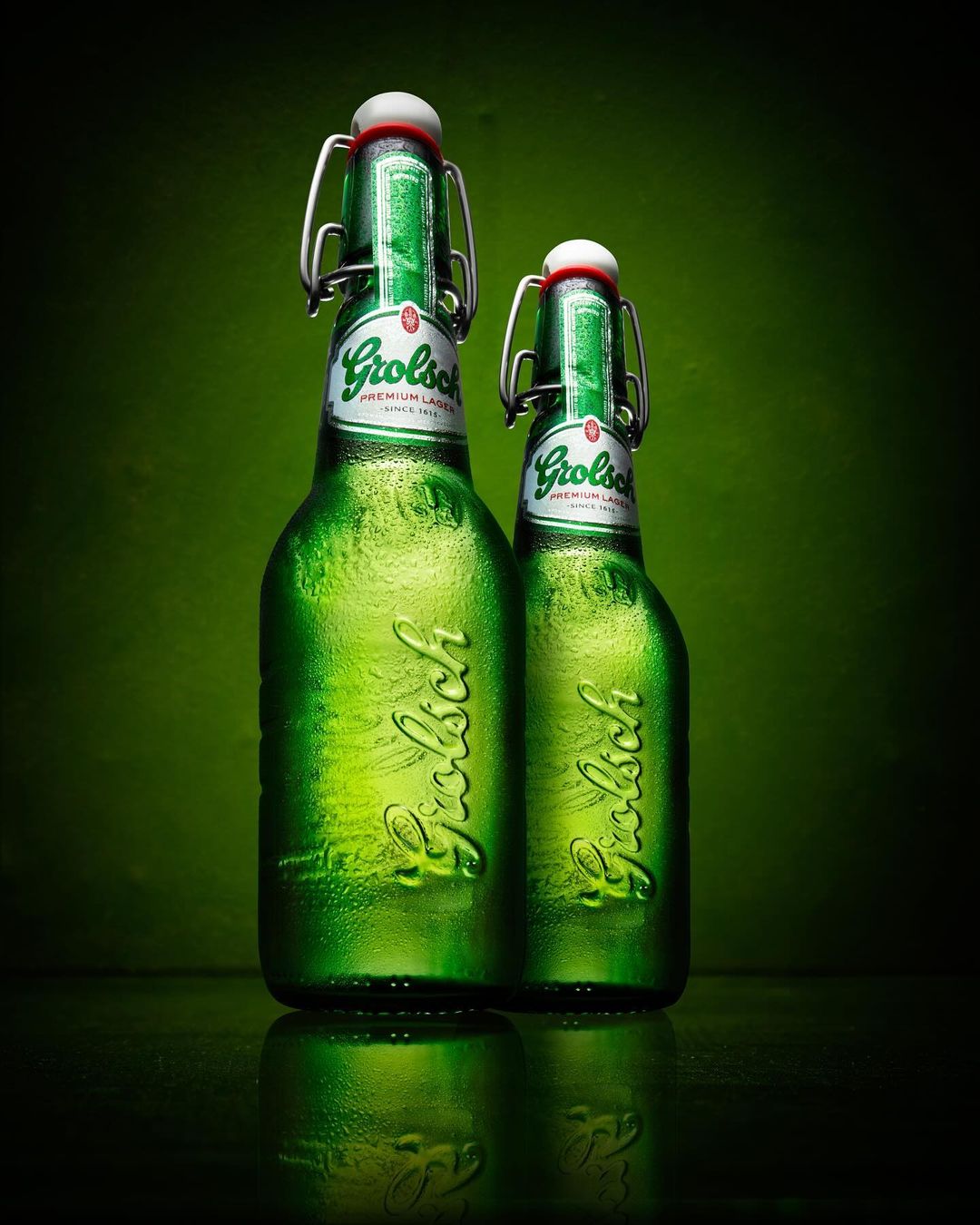 Two green Grolsch beer bottles stand on a dark surface against a green background, capturing a studio shot with focus and lighting.