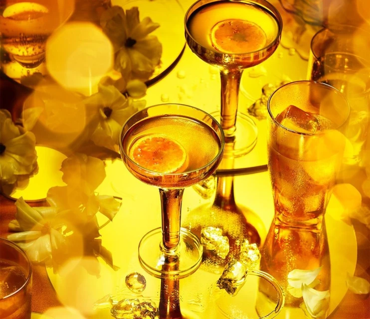 Stemmed coupe glasses filled with amber liquid and citrus slices surrounded by white flowers and ice, all lit with golden lighting.