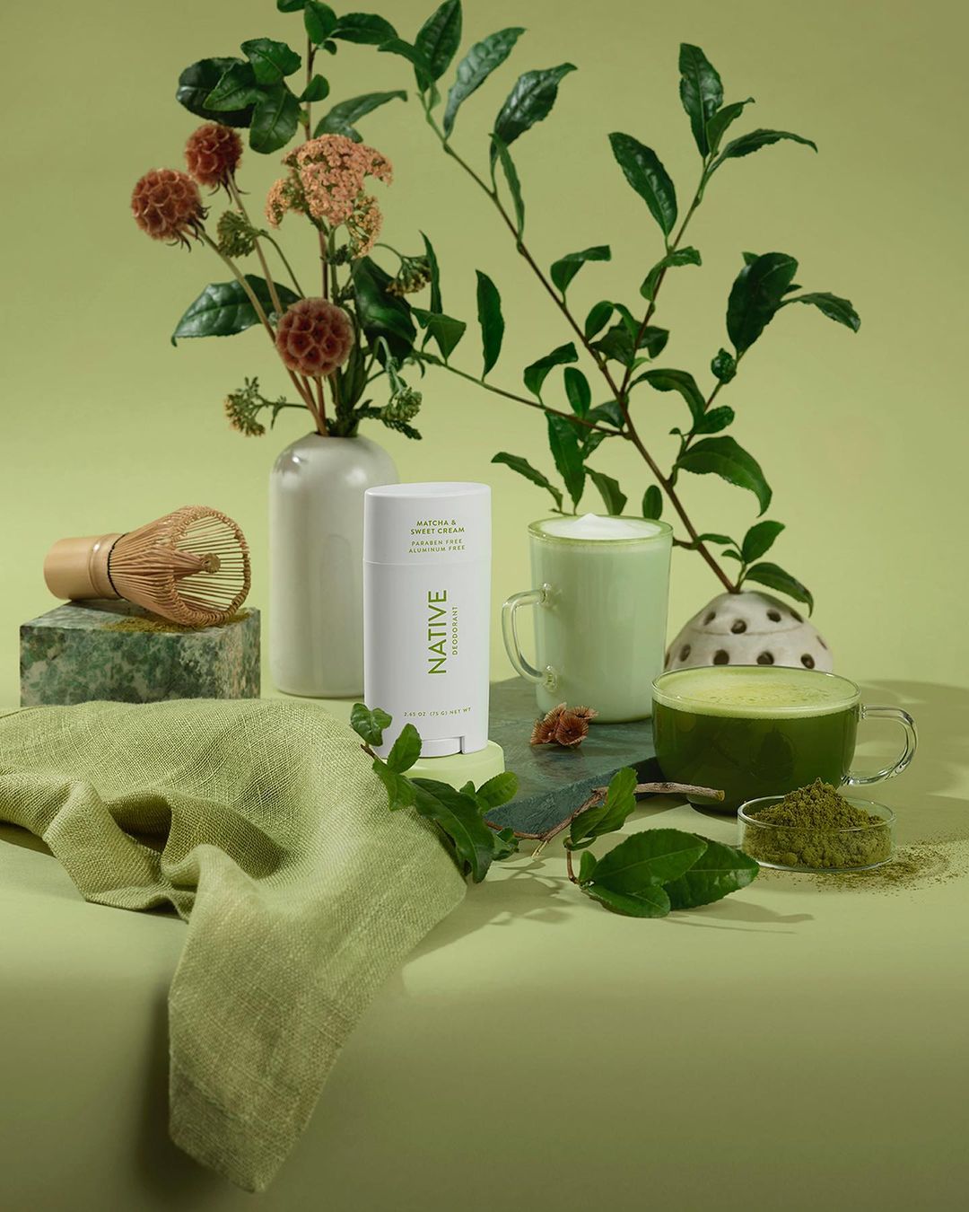 Still life of Native deodorant, matcha tea, and green foliage against a light green background in a studio setting. Product photography.