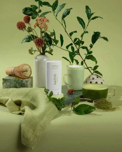 Still life of Native deodorant, matcha tea, and green foliage against a light green background in a studio setting. Product photography.