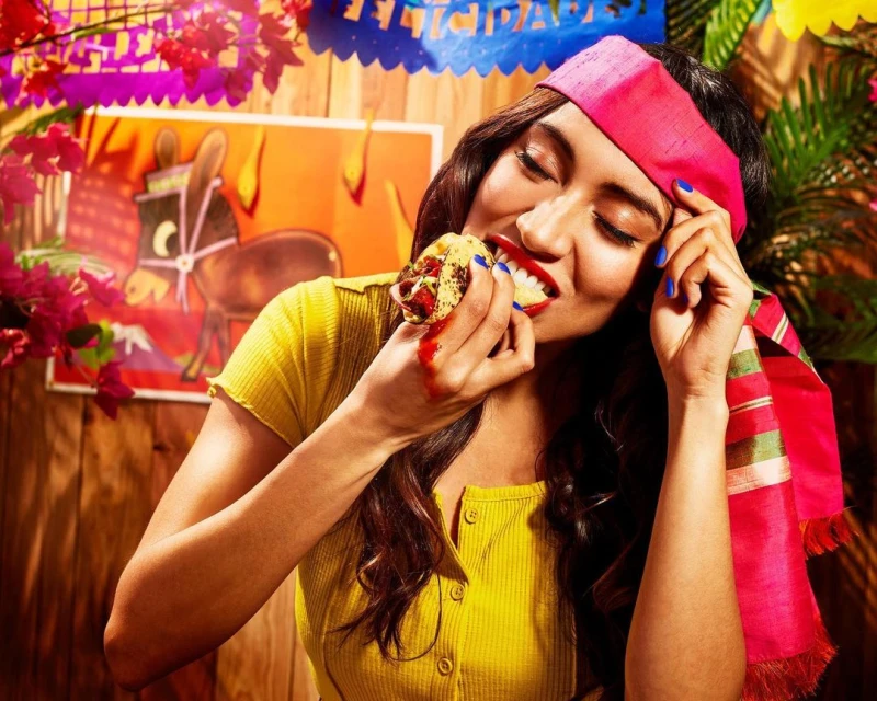 A young woman with a pink headband bites into a taco outdoors, with a colorful painting of a donkey visible in the background.