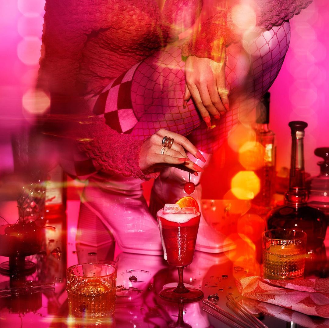 Person's hands adding cherry to red cocktail drink on glass table with liquor bottles, bright pink and red bokeh lighting.