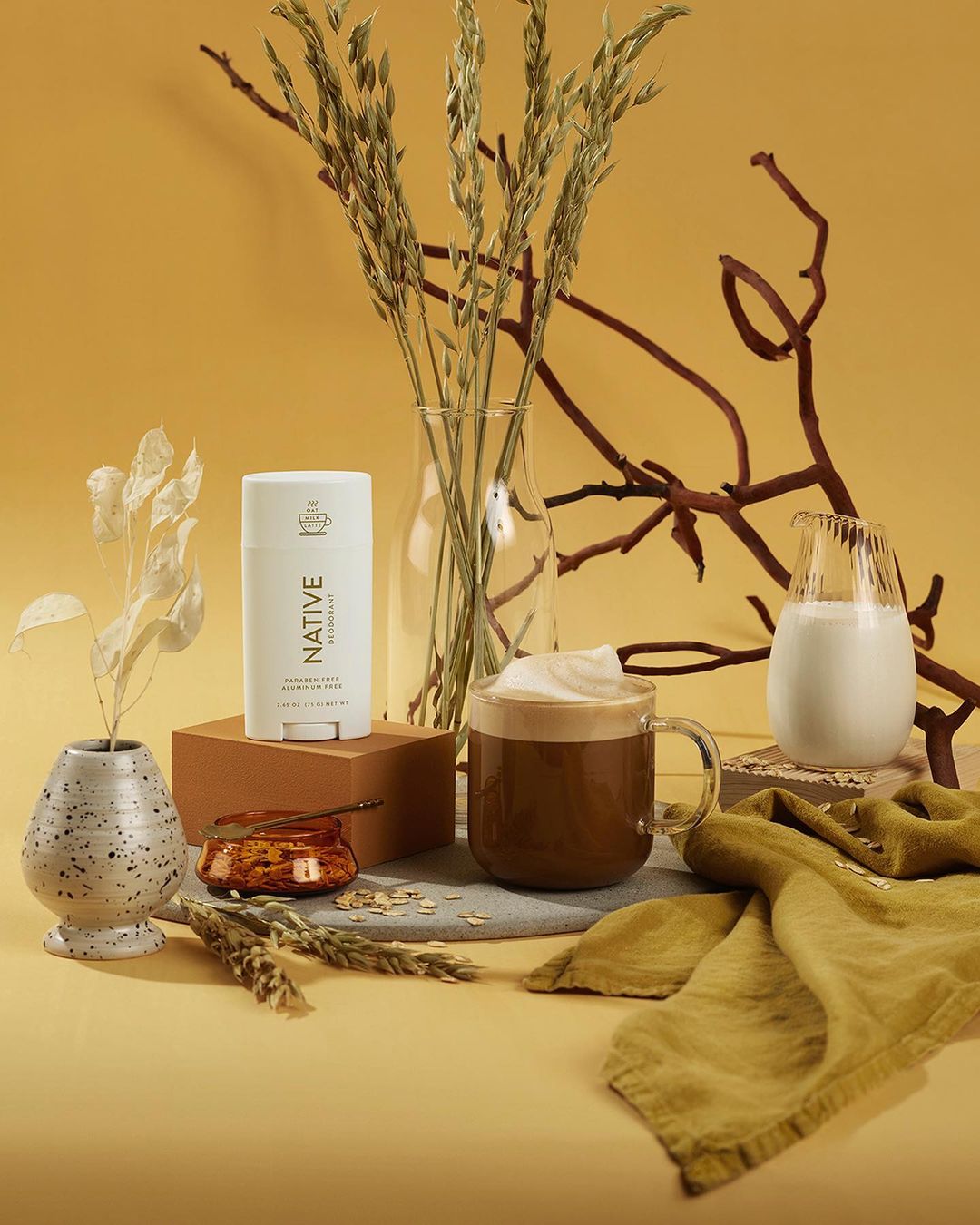 Still life shot of Native deodorant with a glass of coffee, milk, and decorative natural elements on a warm yellow backdrop.