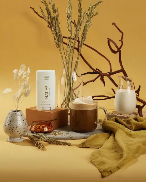 Still life shot of Native deodorant with a glass of coffee, milk, and decorative natural elements on a warm yellow backdrop.