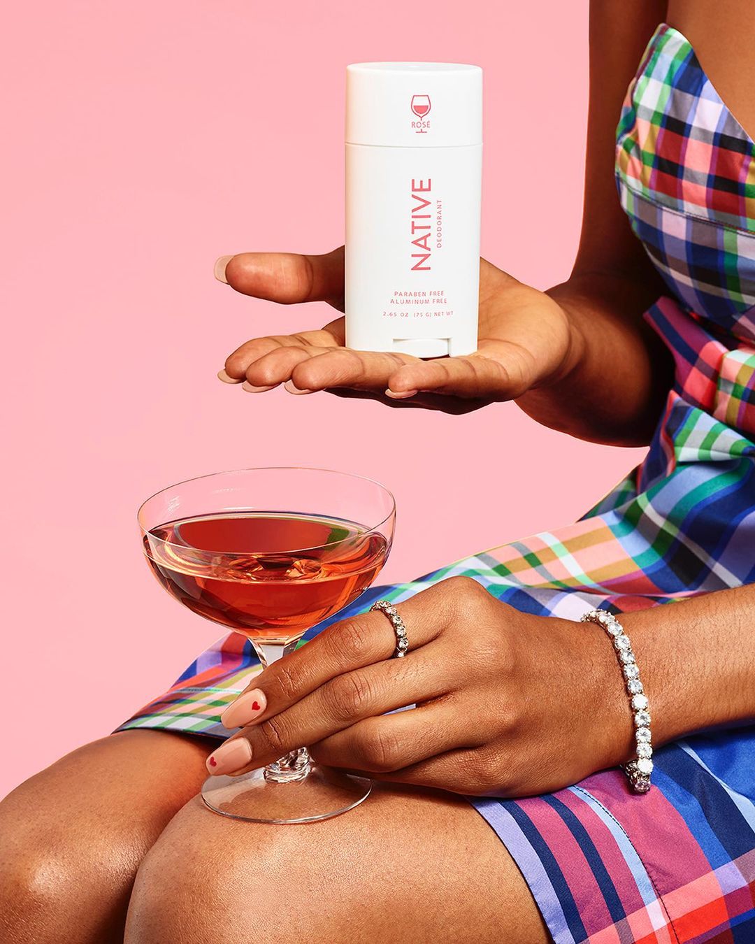Person holding Native brand deodorant and a wine glass against pink backdrop, showcasing beauty and beverage elements.