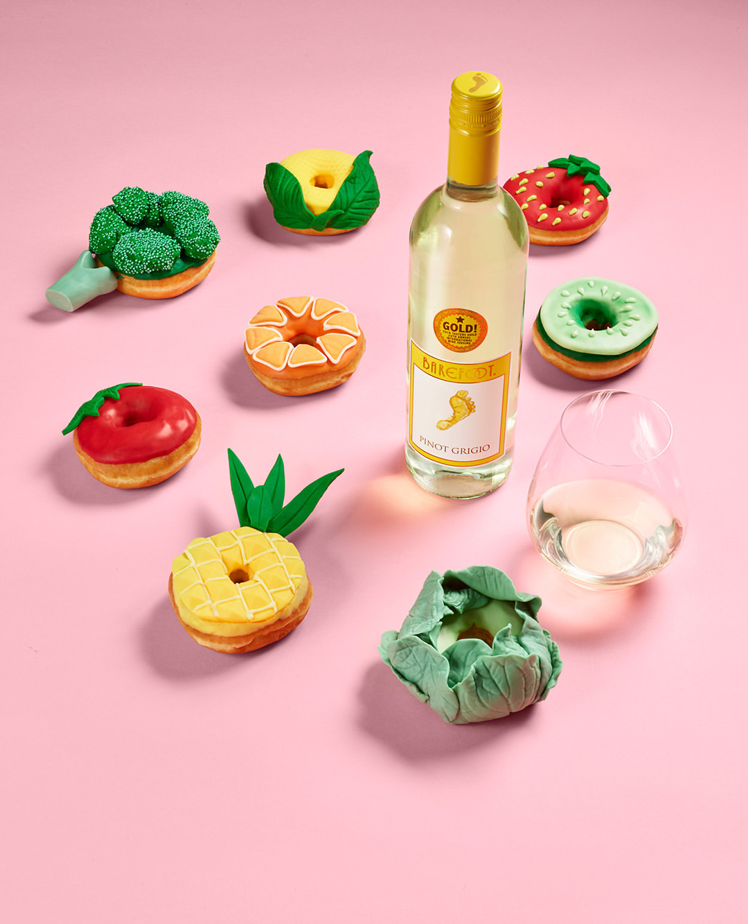 Still life of wine bottle, wine glass and vibrant donuts decorated as vegetables and fruits on a pastel pink background.
