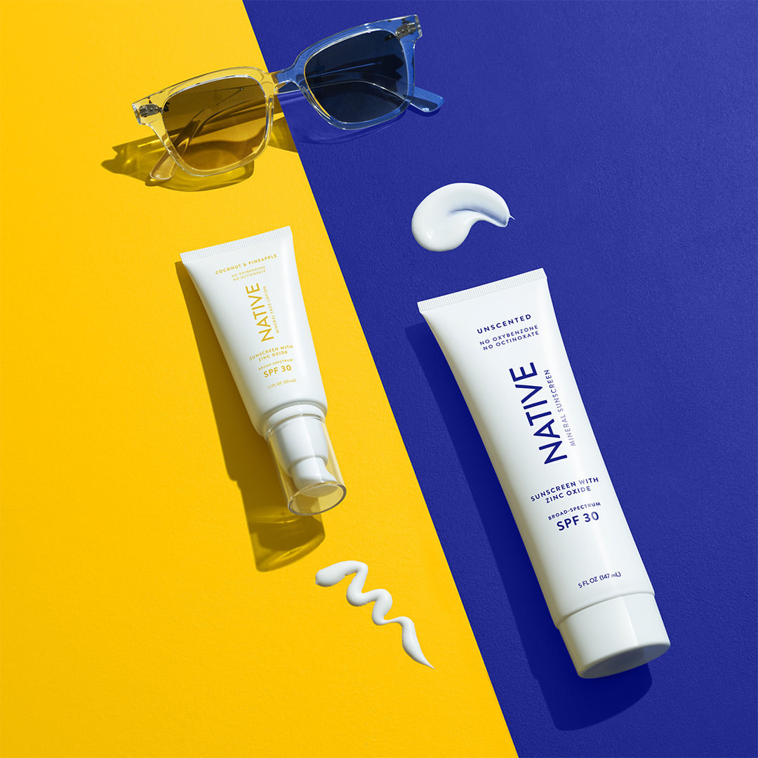 Two tubes of Native sunscreen and pair of sunglasses displayed against a split yellow and blue background create a colorful, summery scene.
