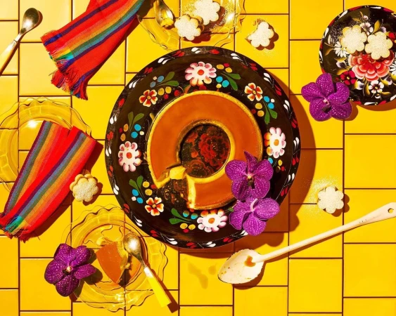 Overhead shot of caramel flan on a decorative plate with floral accents, accompanied by small flower-shaped cakes and purple orchids.