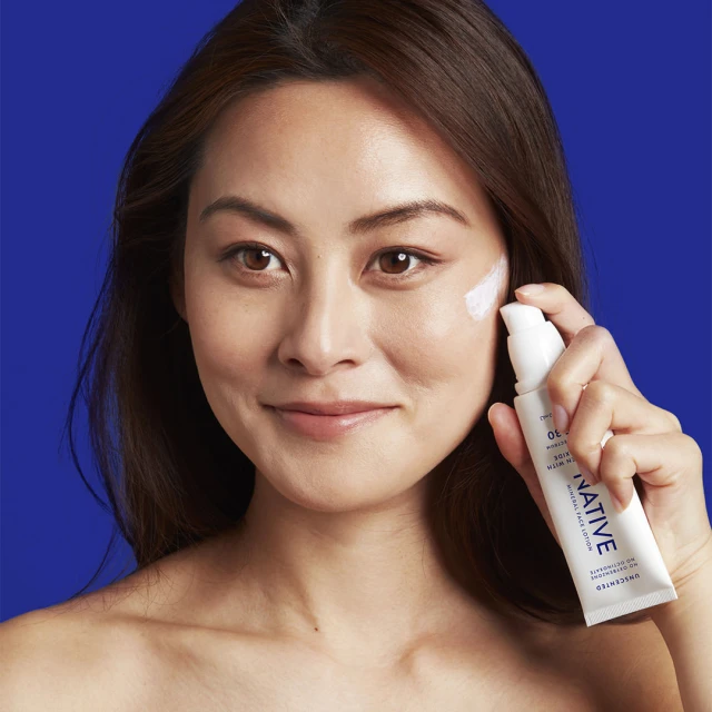 Brunette woman applies white facial cream on cheek in portrait against royal blue backdrop. Tube of lotion held in hand.