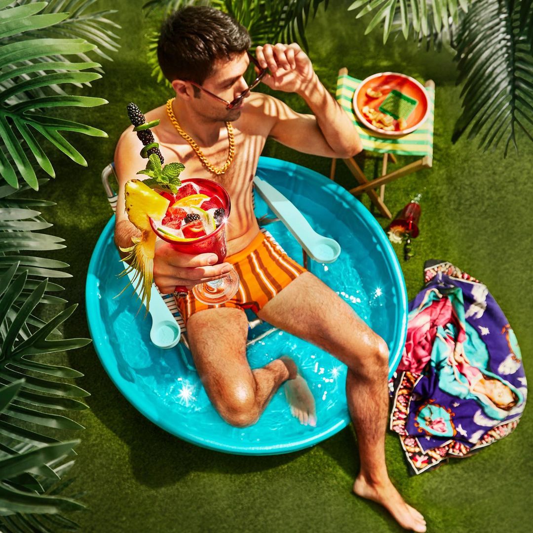Man in swimming pool holding a cocktail and wearing orange swimming trunks, sunglasses and a gold chain, surrounded by green plants.