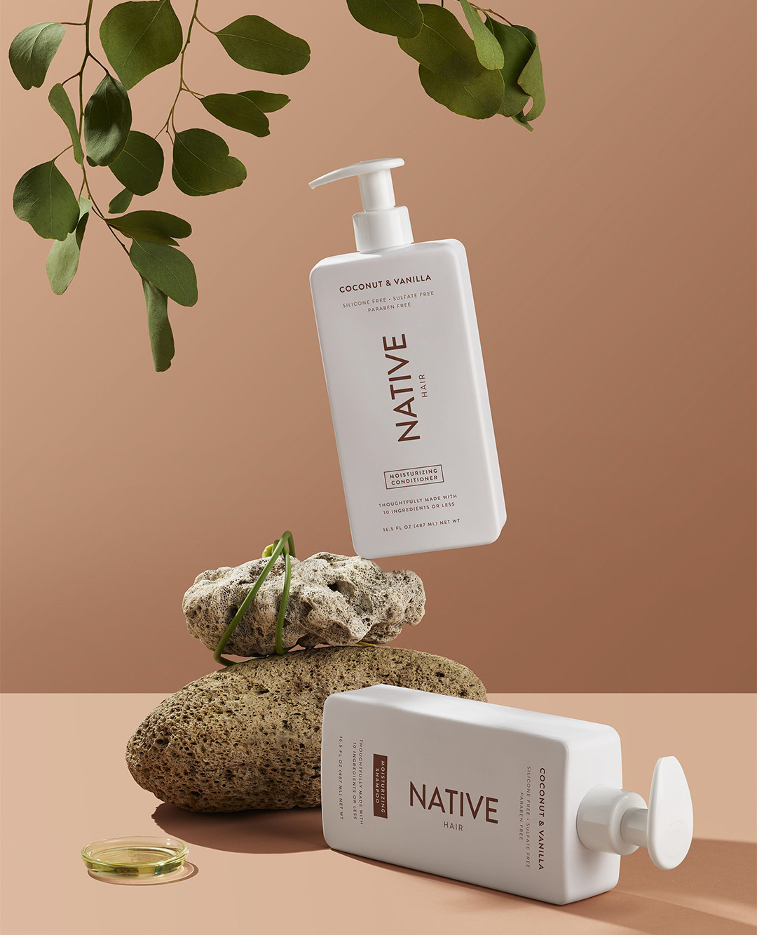 Two bottles of Native Coconut & Vanilla body wash product, arranged next to rocks and eucalyptus leaves against a peach backdrop.