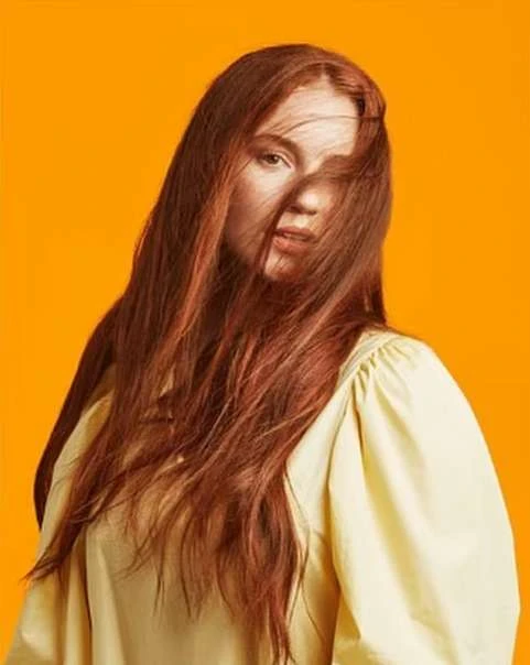 Portrait of a red-haired woman in a light yellow blouse against a solid orange background, hair partially obscuring her face.