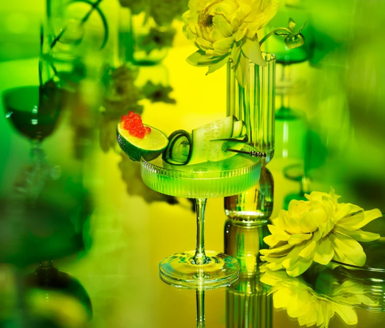 A close-up shot of a green cocktail in a coupe glass garnished with cucumber and roe, with yellow flowers in the background.