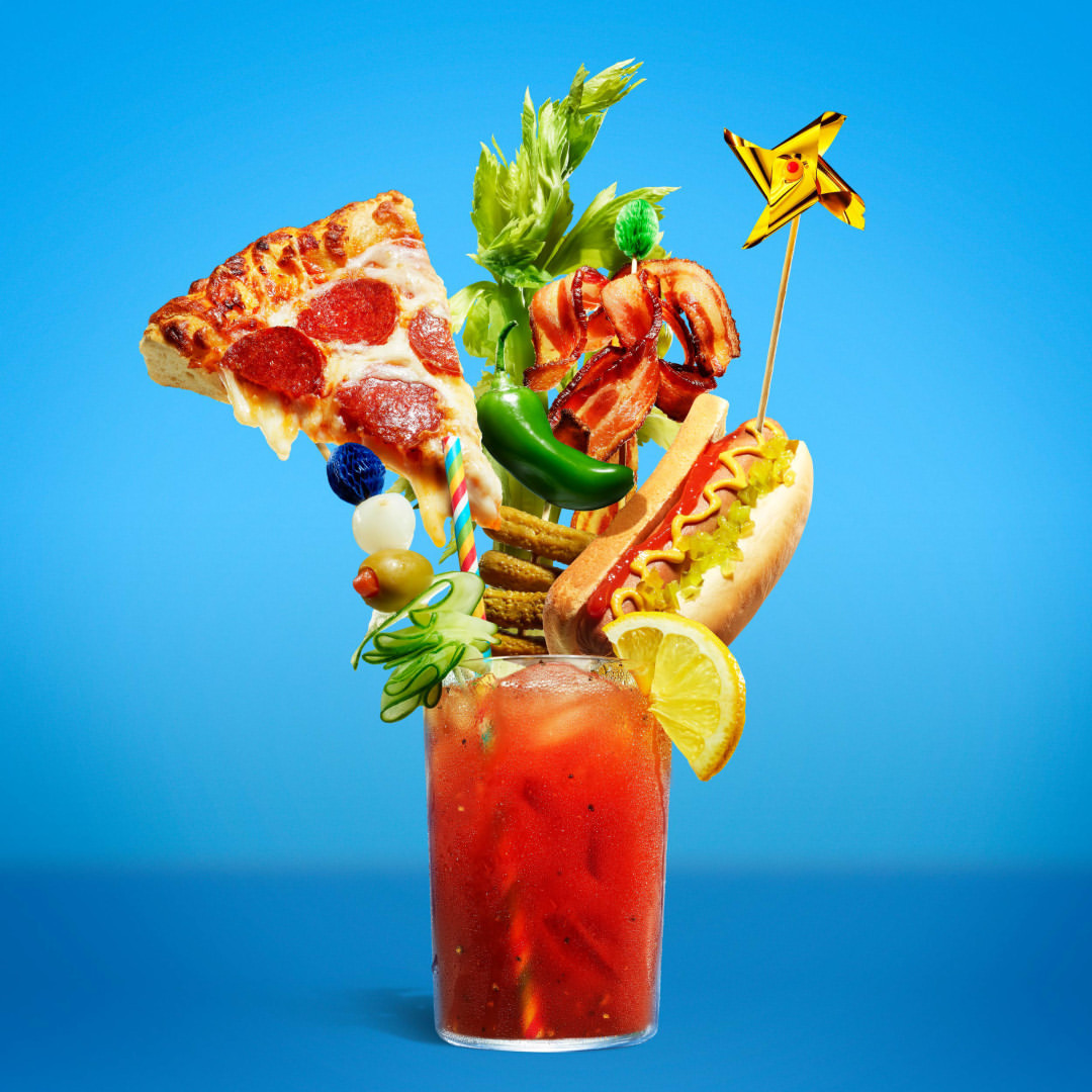 Extravagant Bloody Mary with pizza, hot dog, bacon, jalapeño, celery and lemon slice, against a light blue background in studio shot.