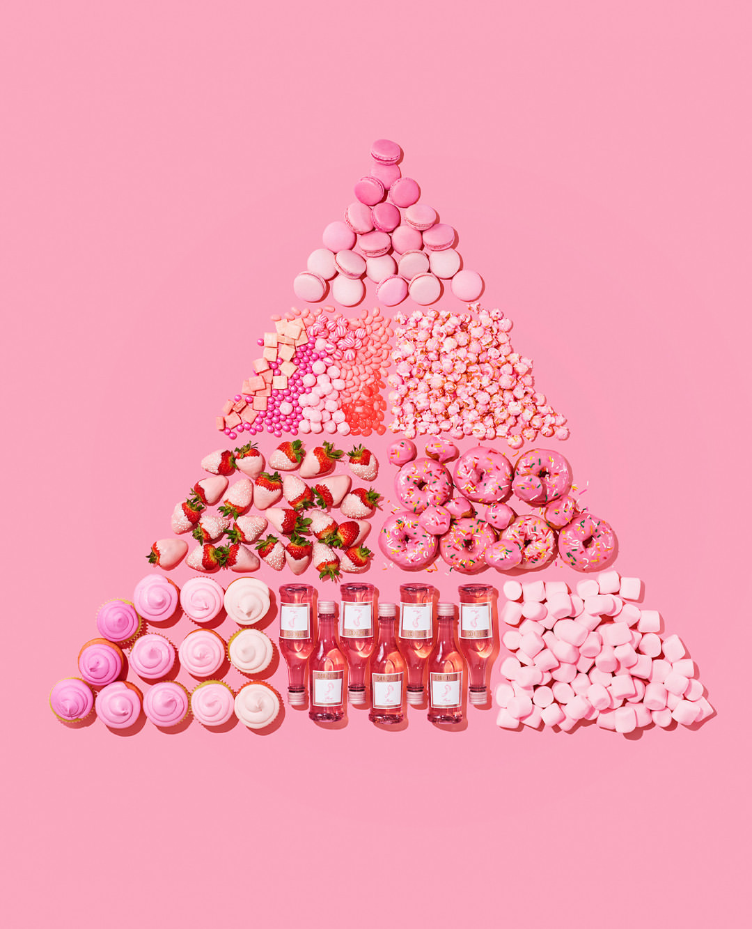 Overhead shot of pink desserts arranged in a triangle shape: macarons, candies, strawberries, doughnuts, cupcakes, mini wine bottles, marshmallows.