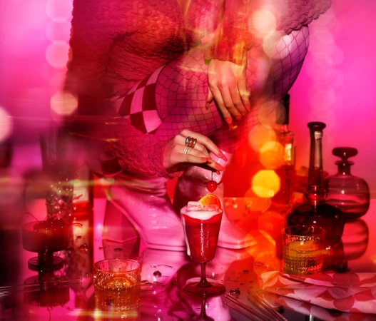 Close-up of a woman in checkered sleeves preparing a cocktail on a reflective surface, bottles and a glass visible in soft lighting.