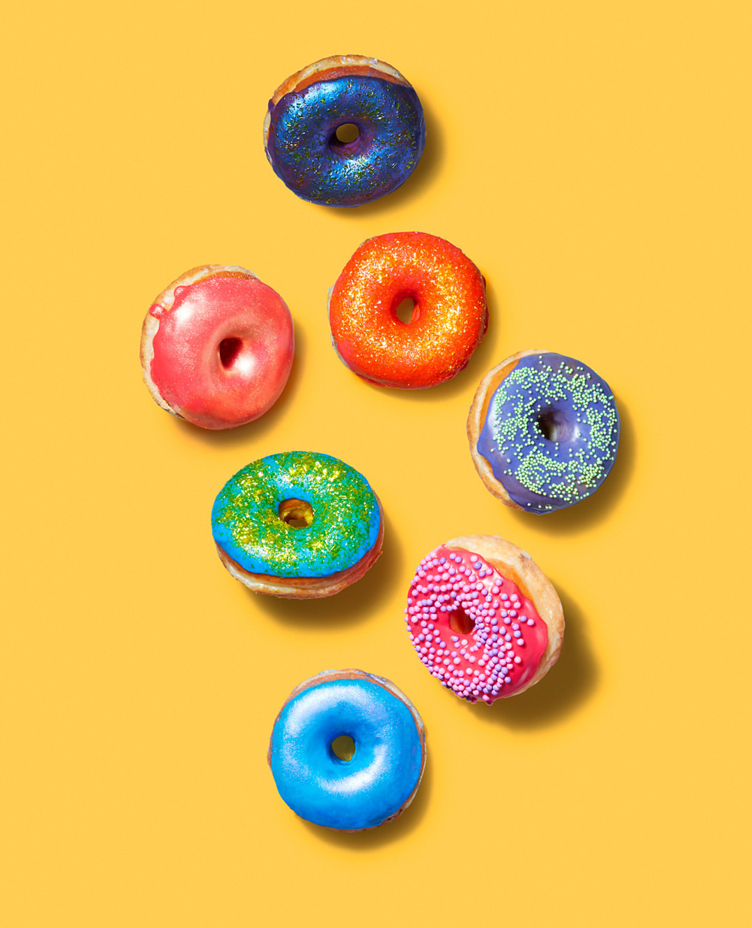 Overhead shot of colorful donuts with various frostings and sprinkles arranged on a bright yellow background in studio lighting.
