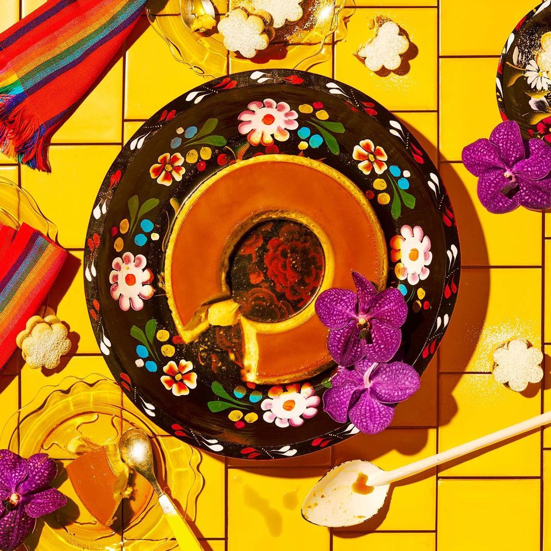 Overhead shot of custard flan dessert on a dark, floral-patterned plate, decorated with purple orchids and flower-shaped cookies.