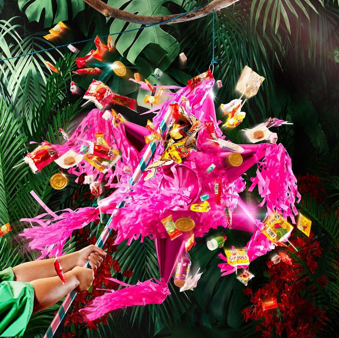 A pink piñata hangs against a backdrop of tropical plants, surrounded by falling candy, coins, and hands holding the rope and stick.